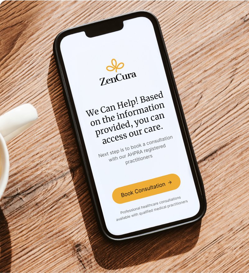 A smartphone on a wooden table displays the ZenCura website, showing a message about accessing care and a yellow "Book Consultation" button. A coffee cup is partially visible on the left.