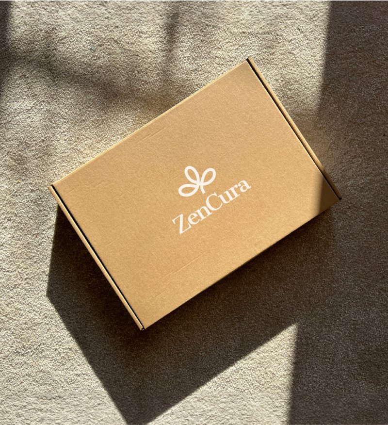 A brown cardboard box with the white ZenCura logo and text is placed on a beige carpet, lit by natural sunlight.