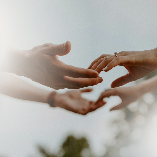 Two pairs of hands reaching towards each other against a blurred outdoor background, with one person wearing a ring.
