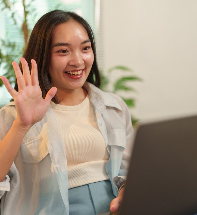 A woman smiles and waves at her laptop during a video call, sitting indoors with plants in the background.