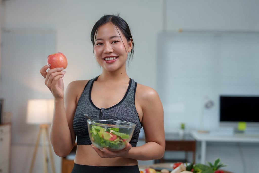 A woman in athletic wear holds a bowl of salad in one hand and an apple in the other, smiling in a modern kitchen—embracing preventive healthcare for long-term wellbeing.